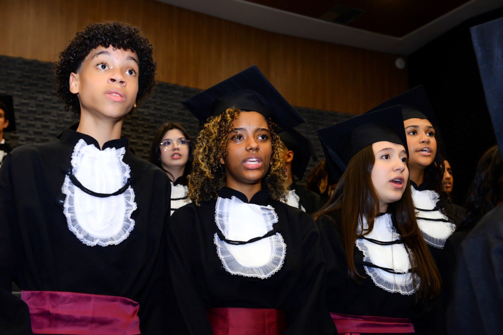 Turma de formandos de beca e capelo cantando durante a colação de grau